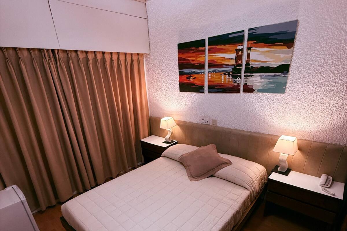 A hotel room with a large bed, two bedside tables with lamps, a painting on the wall, and a window with curtains.