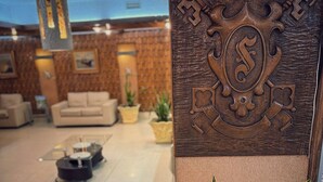 A modern lobby with a large wooden panel featuring a key and a shield emblem, a seating area with a sofa and a coffee table, and potted plants. - Shelton Hotel (Punta del Este)