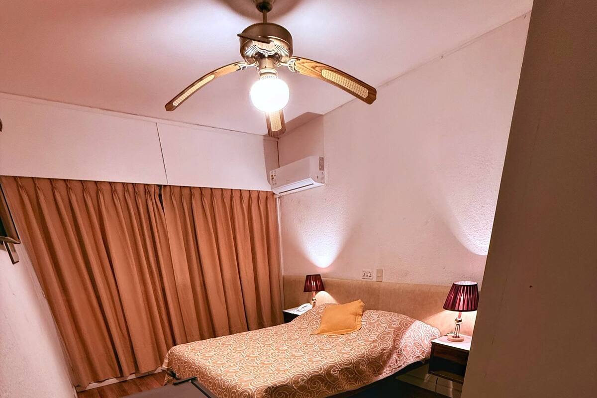 A hotel room with a bed, a ceiling fan, a nightstand with a lamp, and a window with curtains.