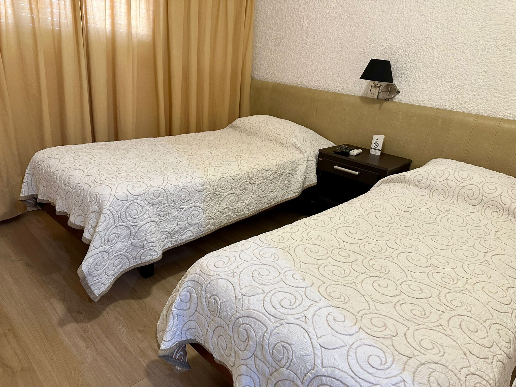Habitación de hotel con dos camas individuales, una mesita de noche con lámpara e iluminación en la pared.