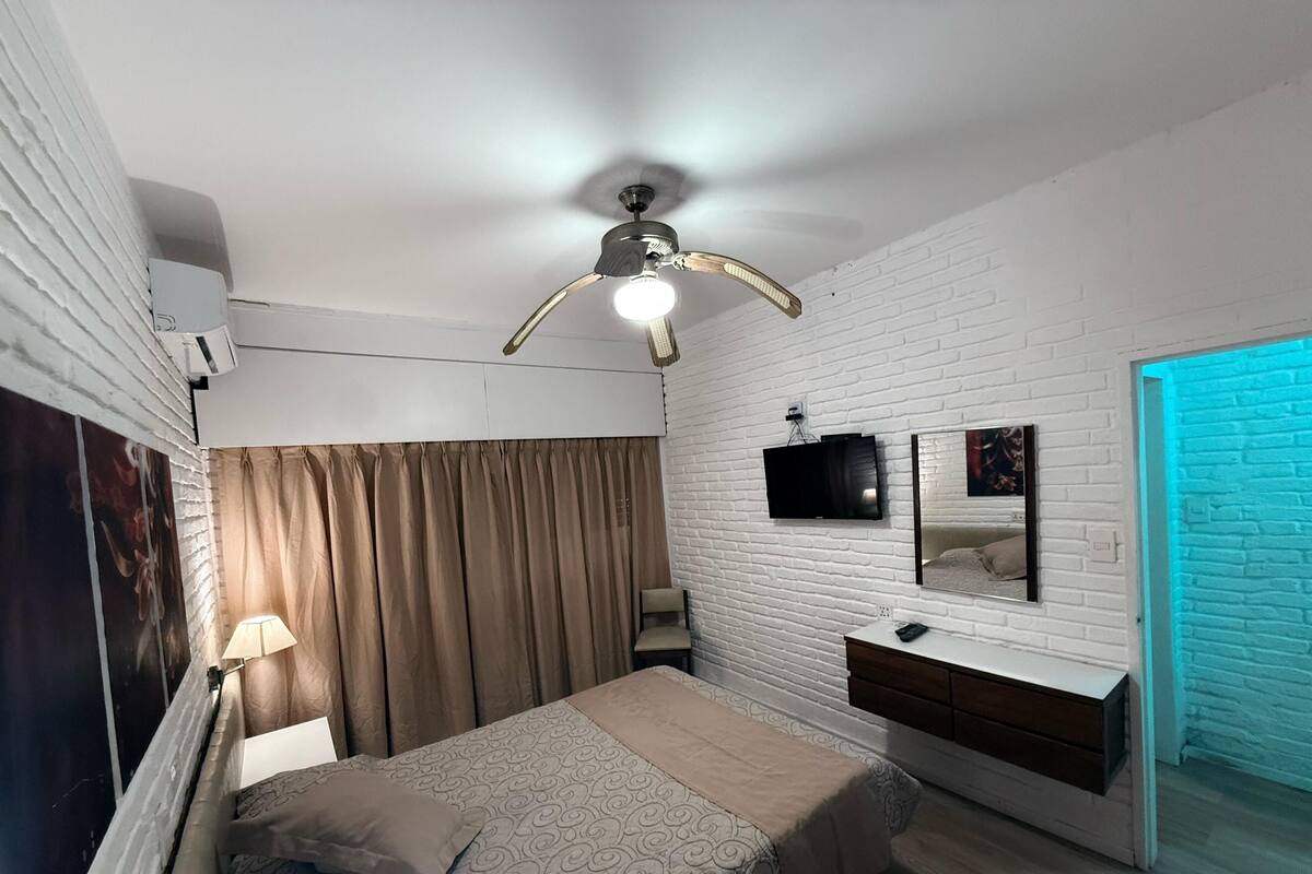 A bedroom with a bed, a ceiling fan, a TV, a mirror, and a blue-lit door.