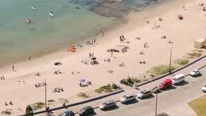 Una playa con agua cristalina, gente tomando el sol y vehículos estacionados a lo largo de la carretera.