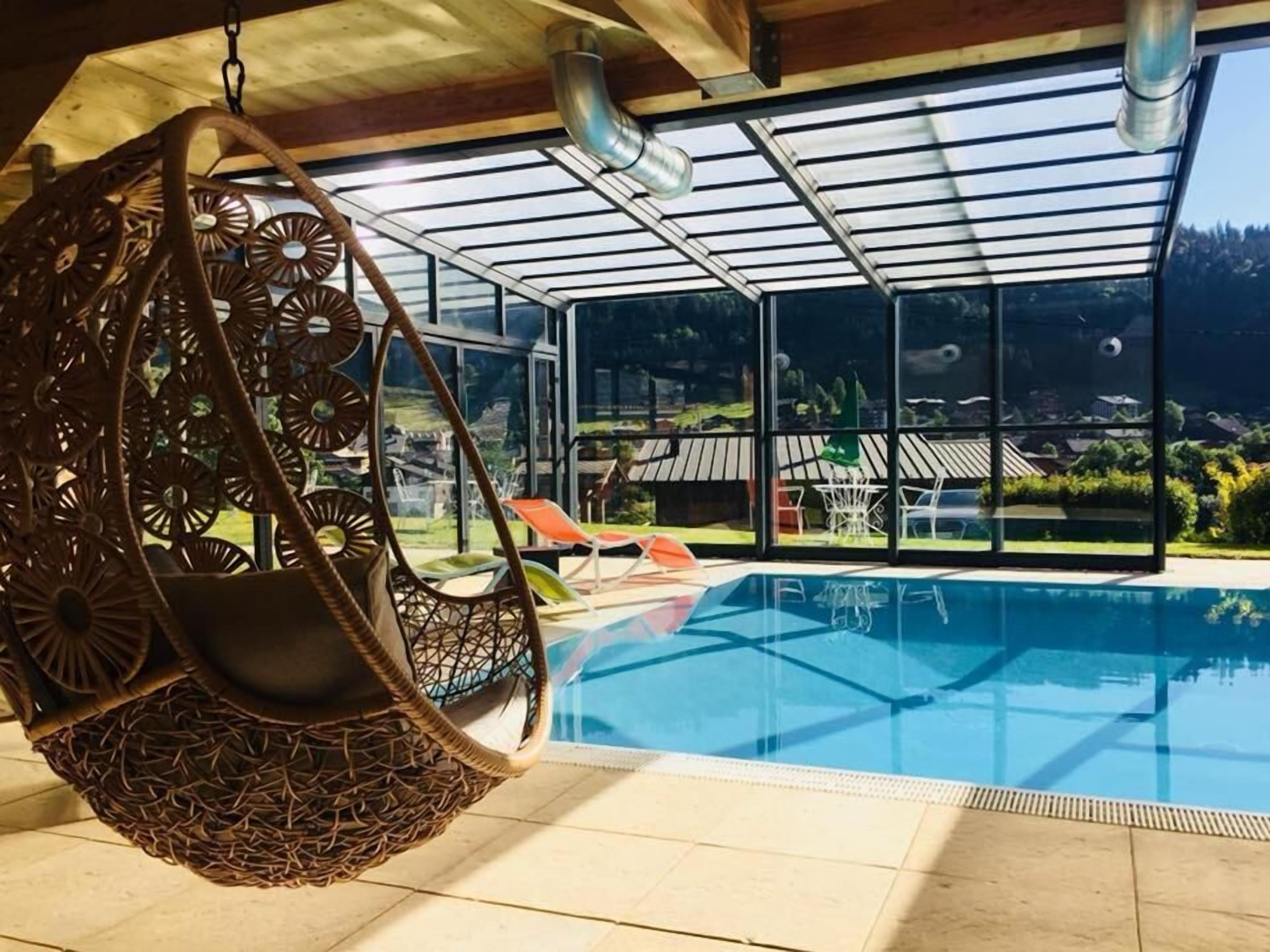 A modern indoor pool area with a hanging chair, clear glass walls, and a view of a residential area.
