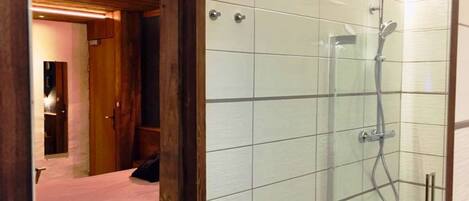 A modern bathroom with a glass shower enclosure, a wooden door frame, and a visible bed in the adjacent room.