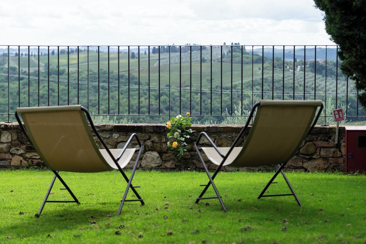 Two outdoor chairs facing a stone wall with a view of a green landscape and a metal fence.