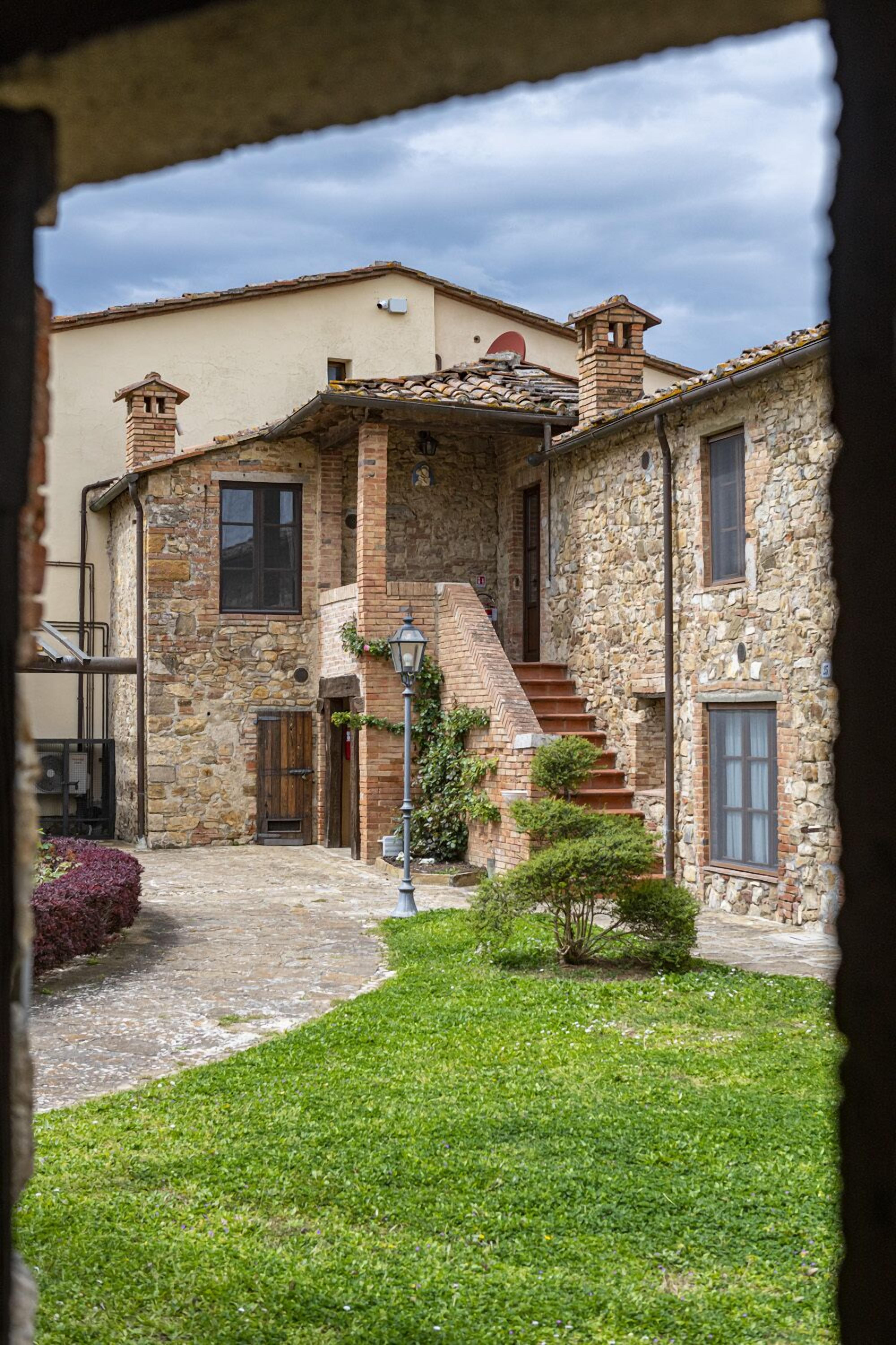 A stone building with a staircase leading to a second floor, surrounded by a small garden and a paved courtyard.