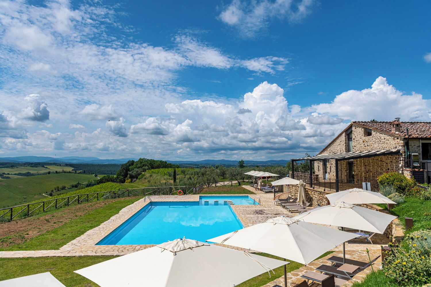Une piscine bordée de pierres, entourée de parasols et de chaises longues, avec en arrière-plan un bâtiment en pierre et un paysage pittoresque.