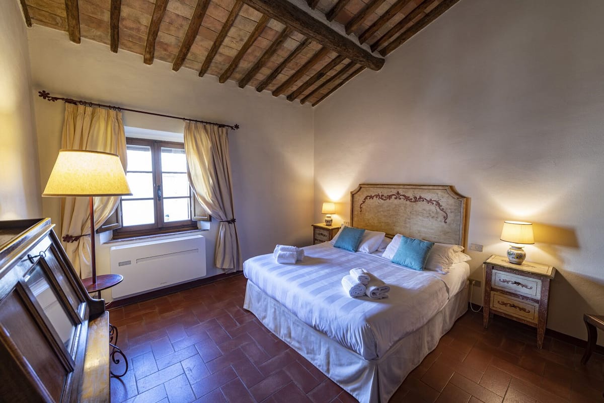 A bedroom with a bed, two bedside tables with lamps, a window with curtains, a radiator, and a wooden ceiling.