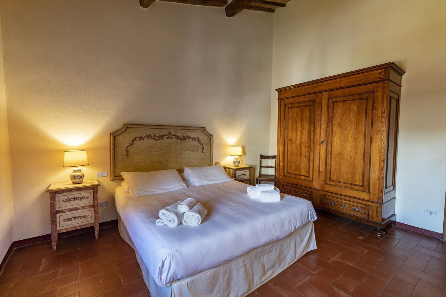A bedroom with a large bed, two bedside tables with lamps, a wooden wardrobe, and a tiled floor.