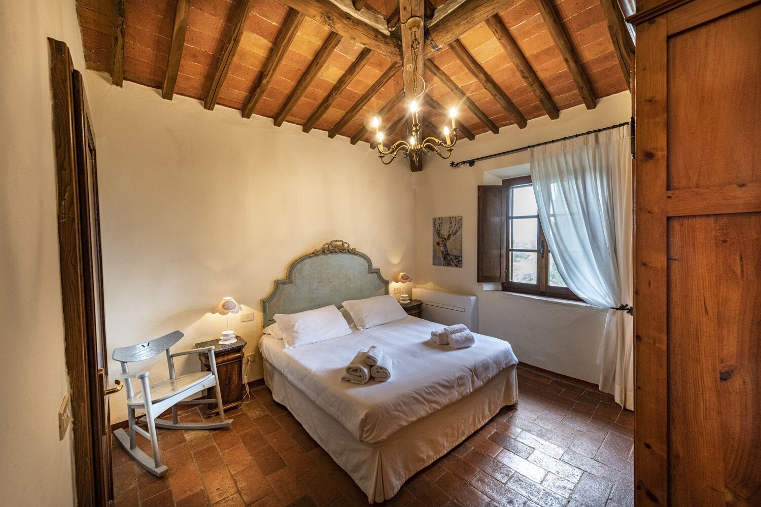Un dormitorio con techo de madera, una cama con ropa blanca, una mesita de noche con lámpara y una ventana con cortinas.
