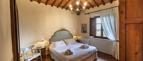Un dormitorio con techo de madera, una cama con ropa blanca, una mesita de noche con lámpara y una ventana con cortinas.