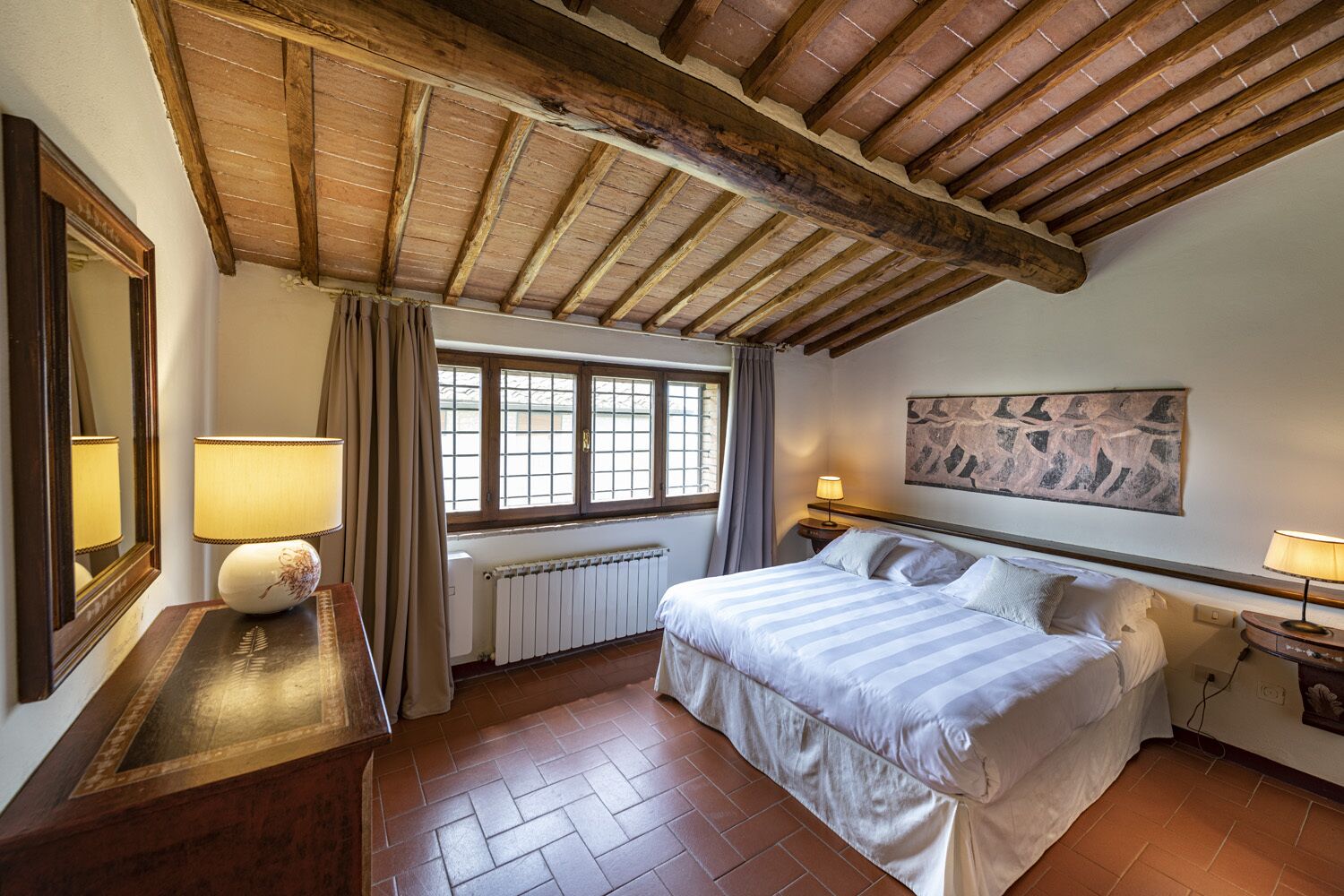A bedroom with a wooden ceiling, a large bed, a window with curtains, a radiator, and a painting on the wall.