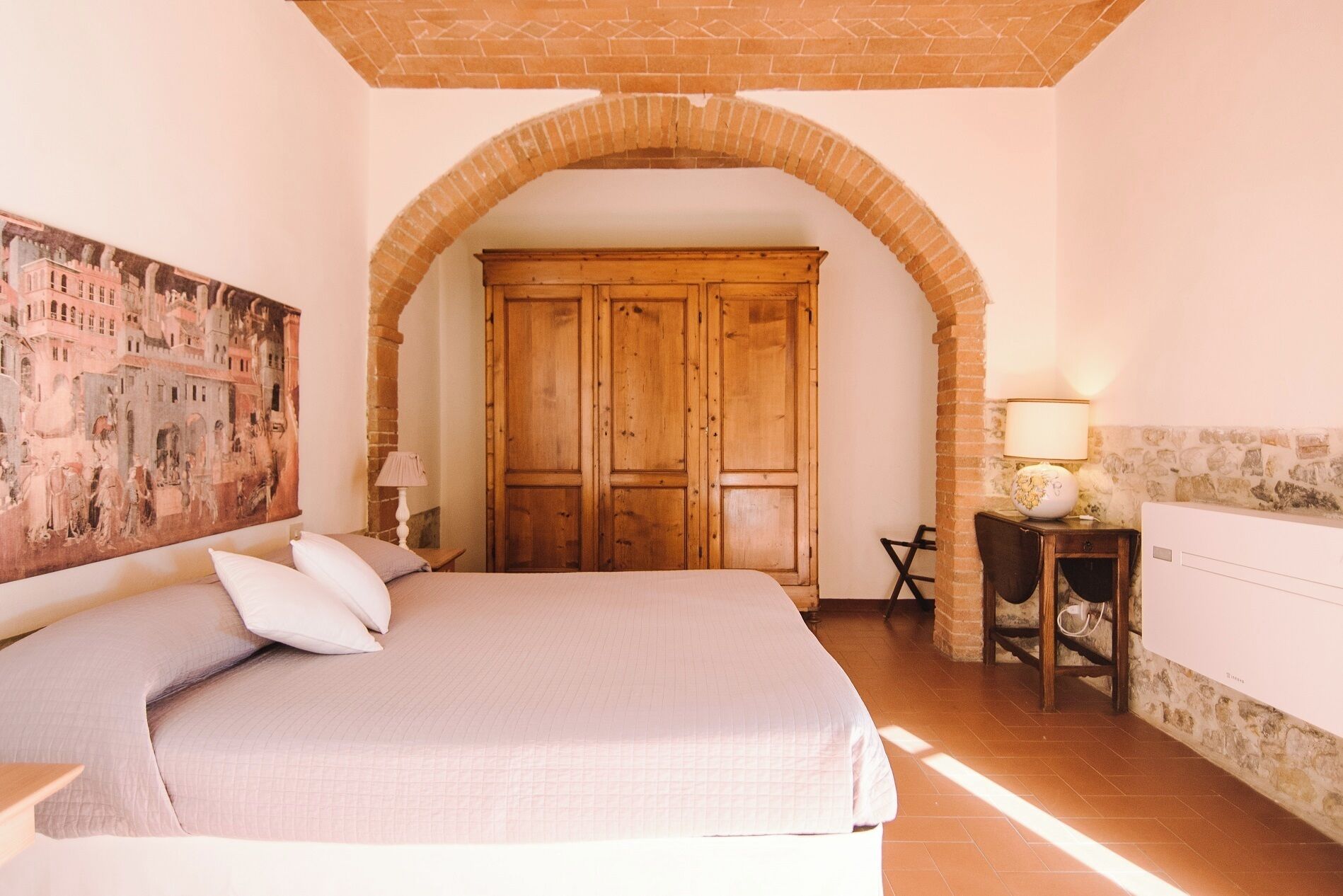 A bedroom with a large bed, an arched doorway, a wooden wardrobe, a desk with a lamp, and a wall painting.