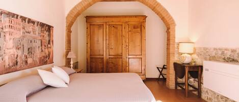 A bedroom with a large bed, an arched doorway, a wooden wardrobe, a desk with a lamp, and a wall painting.