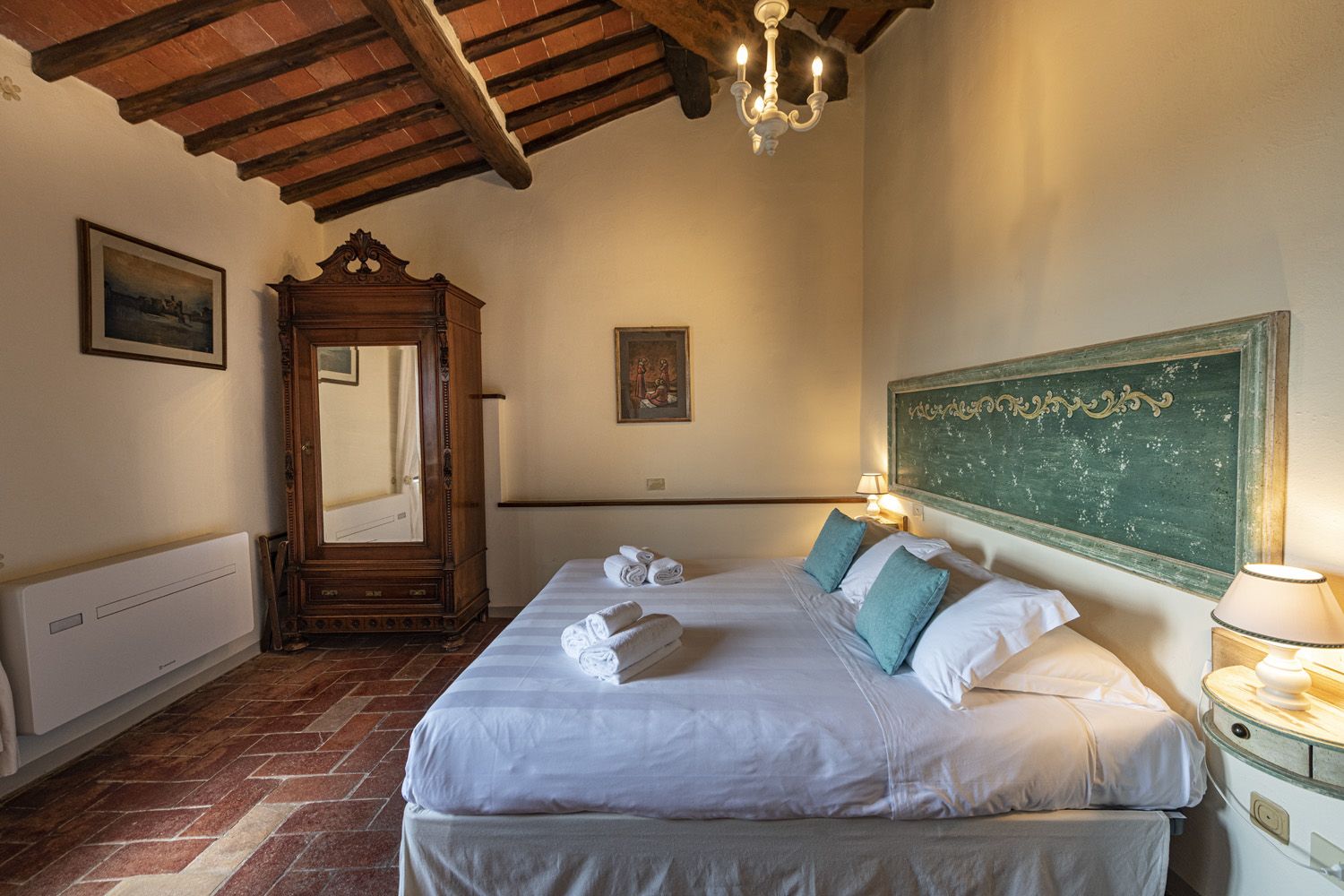 A hotel room with a bed, a wooden wardrobe, a mirror, a wall-mounted picture, a radiator, and a green chalkboard.