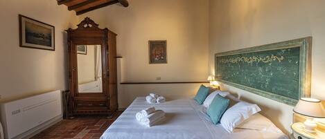 A hotel room with a bed, a wooden wardrobe, a mirror, a wall-mounted picture, a radiator, and a green chalkboard.