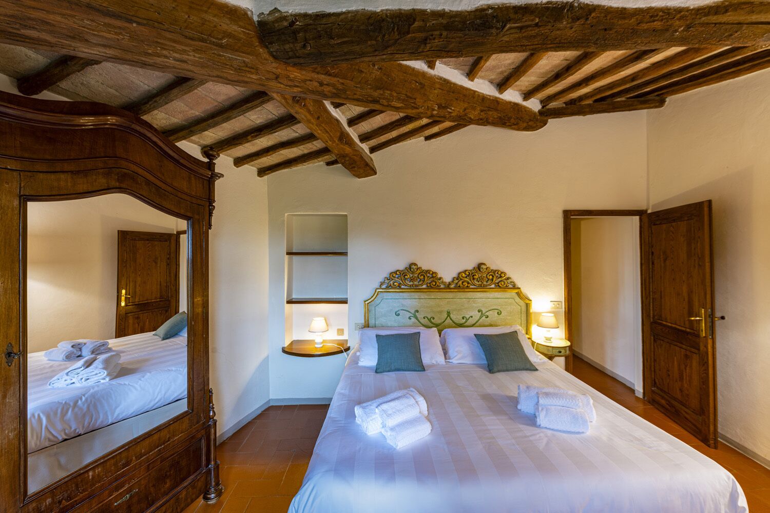 A hotel room with a large bed, wooden ceiling beams, a mirror, and a bedside table with a lamp.