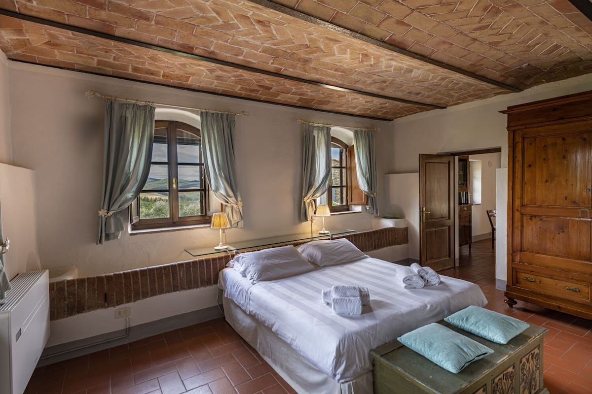 A bedroom with a brick ceiling, a bed with white linens, two windows with curtains, a wooden wardrobe, and a radiator.