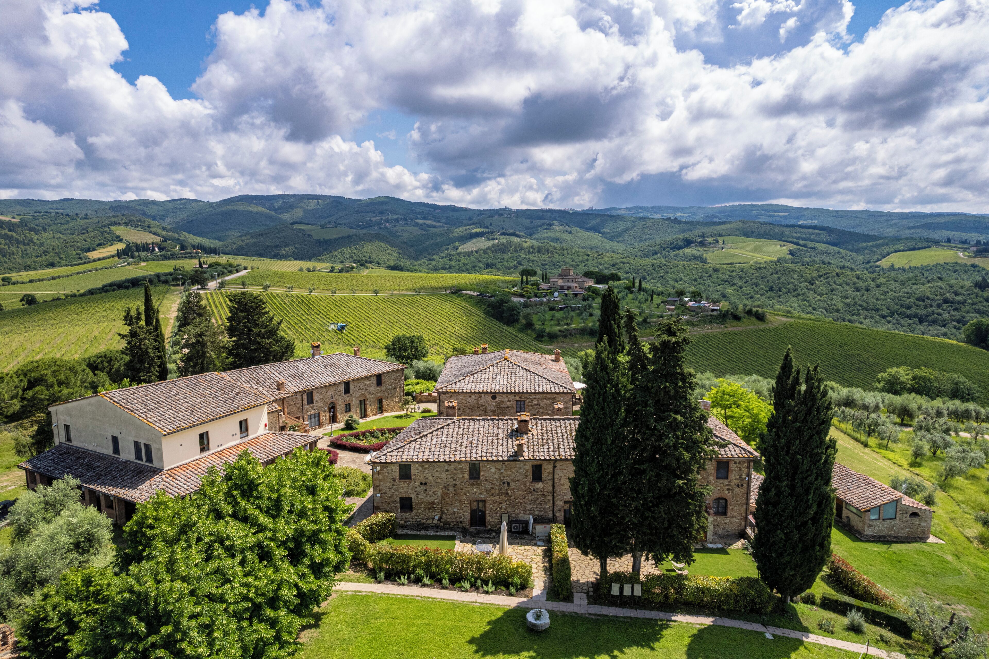 A stone building complex with multiple structures, surrounded by lush green fields and trees, under a partly cloudy sky.