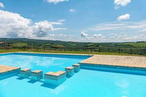 A clear blue pool with stone steps and a stone deck, surrounded by a fence with a view of green hills and a clear sky.