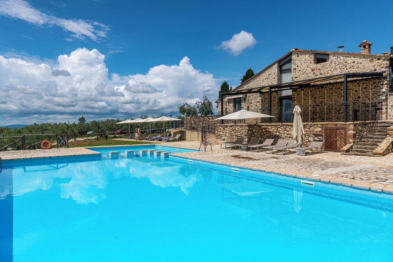 Un edificio de piedra con piscina, sillas de descanso y sombrillas.