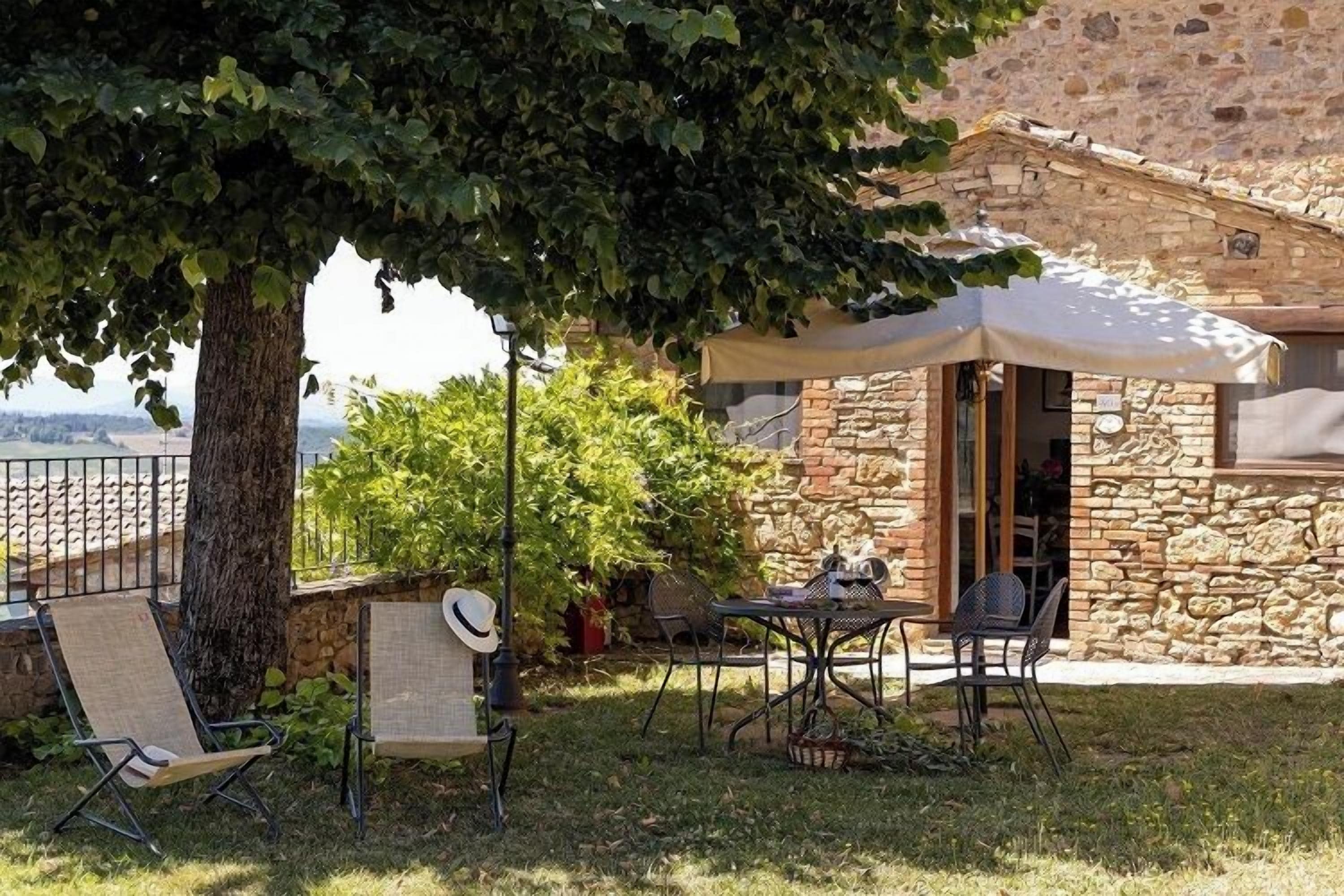 A stone building with a covered outdoor seating area, a tree providing shade, and a view of the landscape.