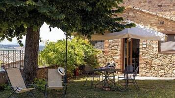 A stone building with a covered outdoor seating area, a tree providing shade, and a view of the landscape.