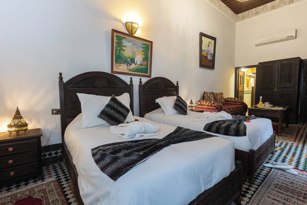 A double bed with white linens, two pillows, and a blanket. There is a nightstand with a lamp and a wall-mounted air conditioner.