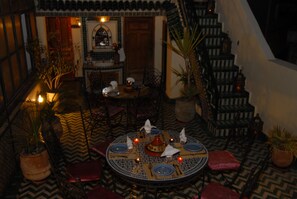 A dining area with a round table set for four, surrounded by chairs. There are potted plants and a staircase in the background. - Riad Ghita (Fes)