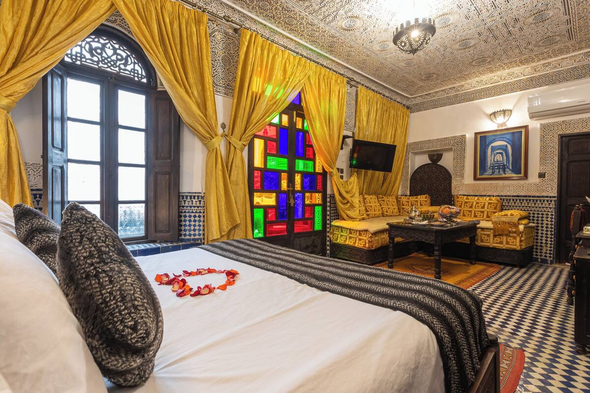 A room with a bed, a sofa, a table, and a decorative window with stained glass.