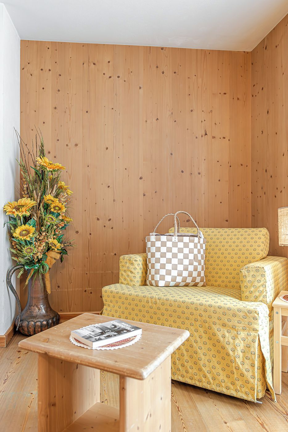 Ein Zimmer mit Holzwänden, einem gelben Sofa, einer karierten Tasche, einer Vase mit Sonnenblumen und einem kleinen Holztisch.