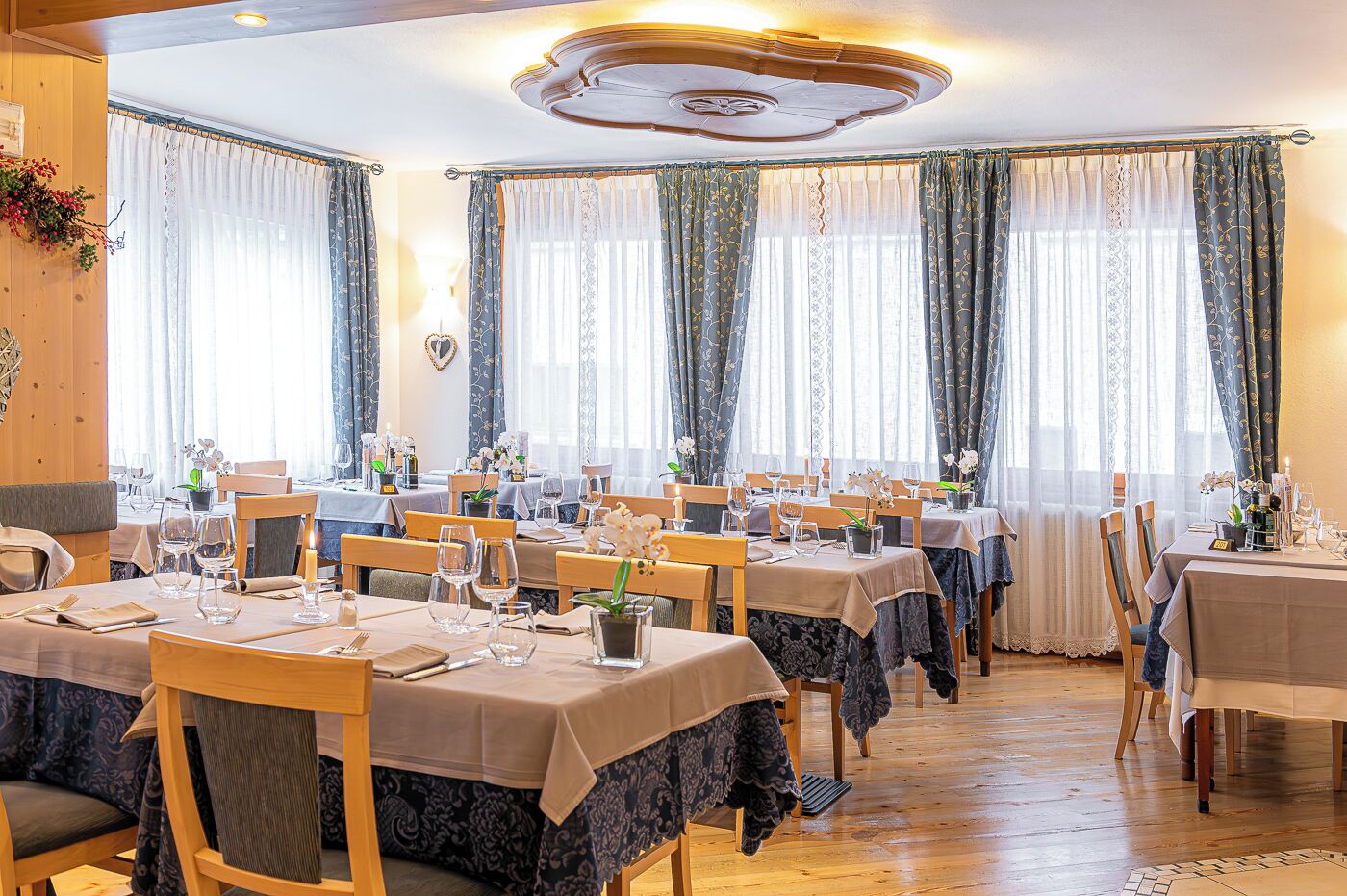 A dining area with multiple tables set for service, large windows with curtains, and a wooden ceiling.