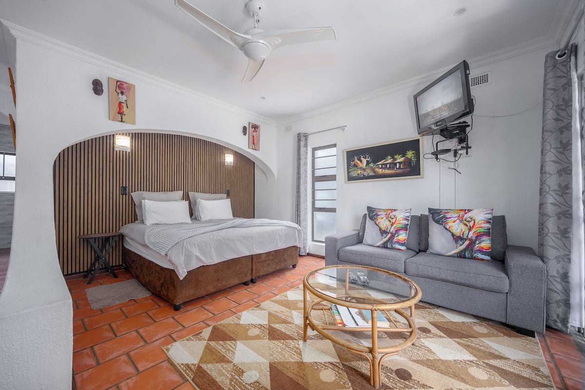 A modern bedroom with a bed, a grey sofa, a TV mounted on the wall, and a ceiling fan.