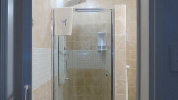 Una ducha con cerramiento de vidrio, baldosas color beige, una toalla colgada en la puerta y un enchufe visible en la pared.