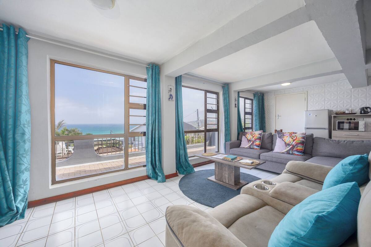 A spacious living room with a large sliding glass door leading to a balcony, a grey sofa with teal cushions, and a small kitchen area with a refrigerator and microwave.
