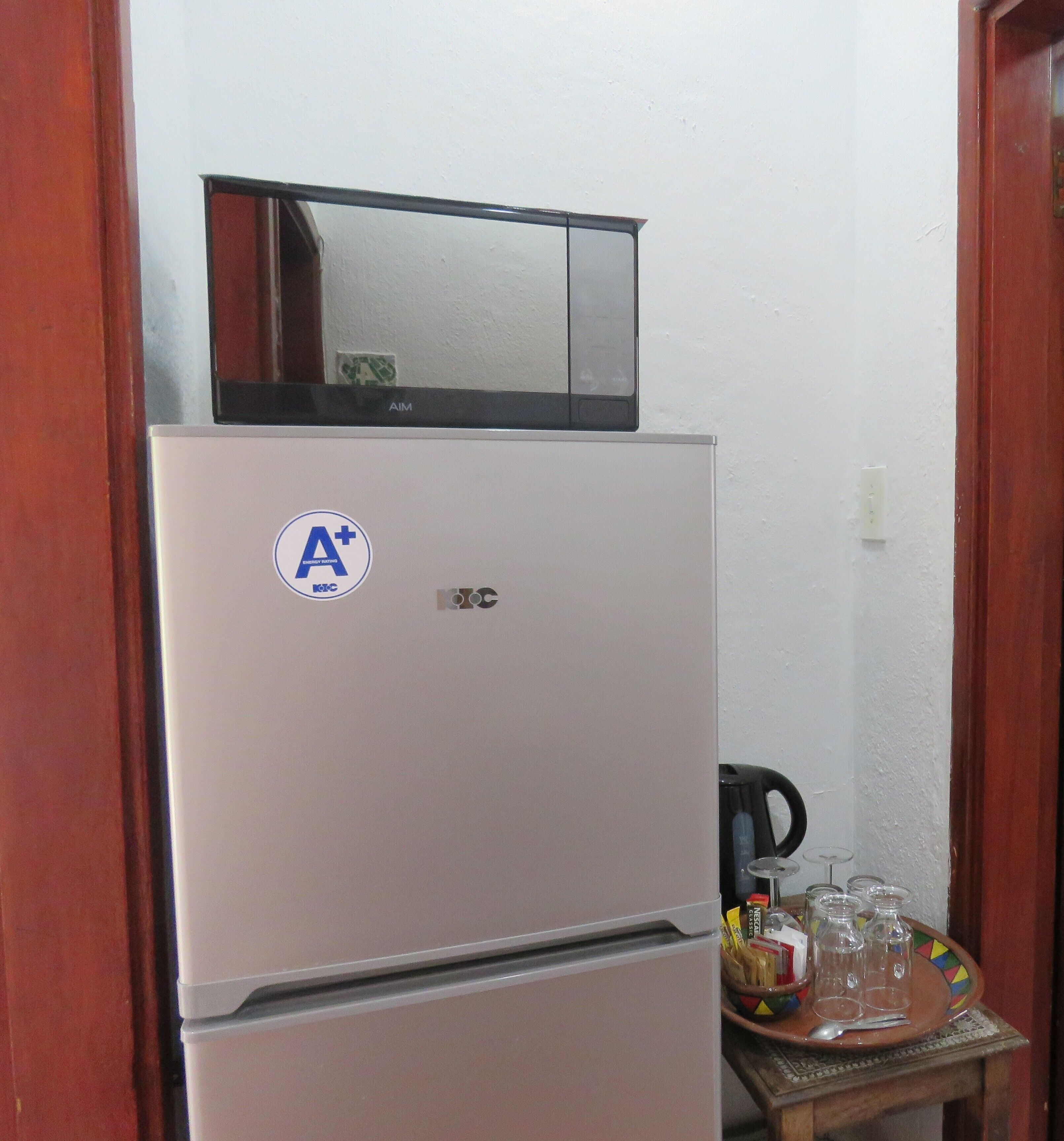 A white refrigerator with a black top unit, a sticker with "A+" and "KFC" logos, and a small table with a kettle and glasses.