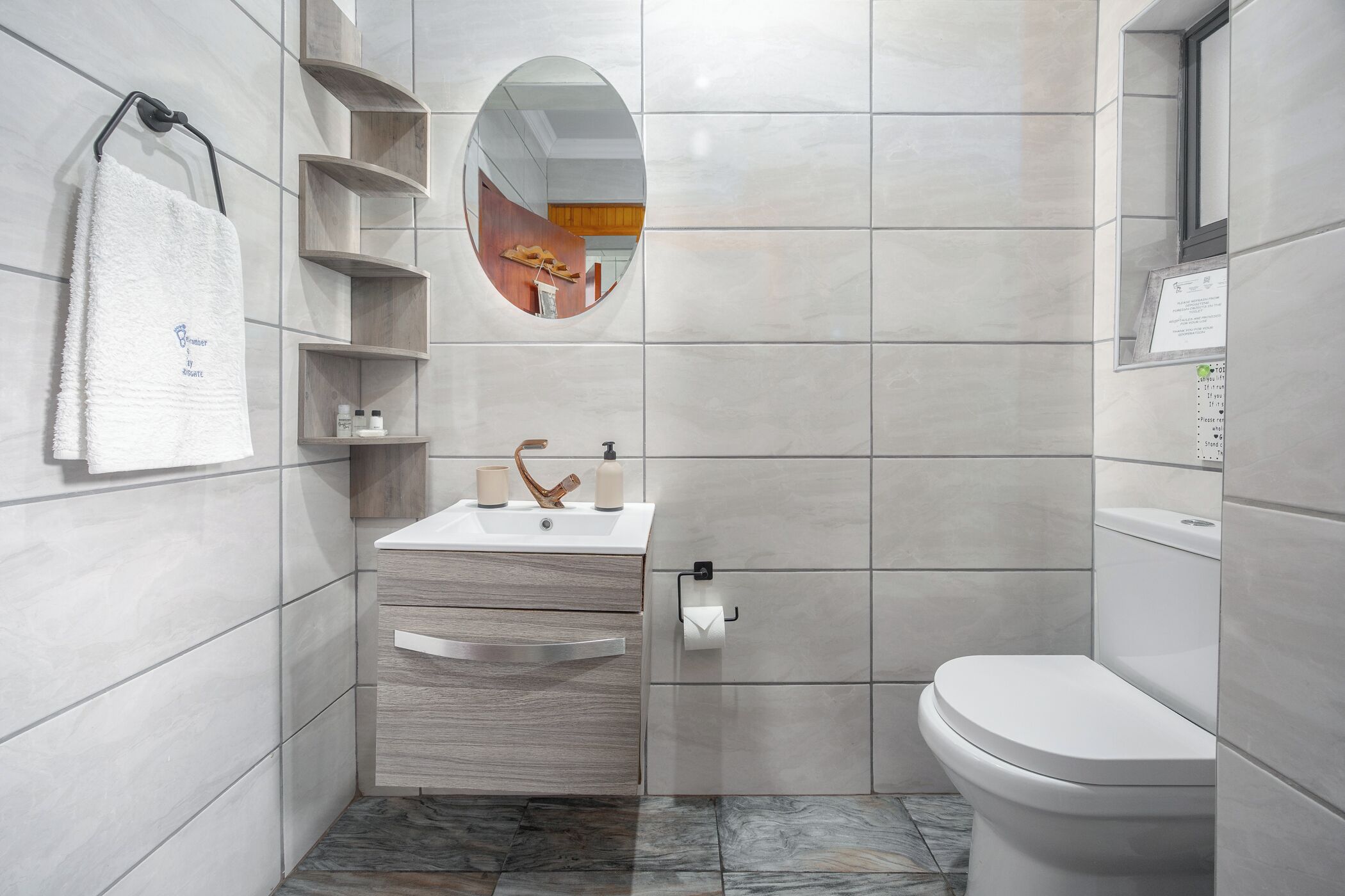 A modern bathroom with a white sink, wooden vanity, and a toilet. There is a towel hanging on the wall and a mirror above the sink.