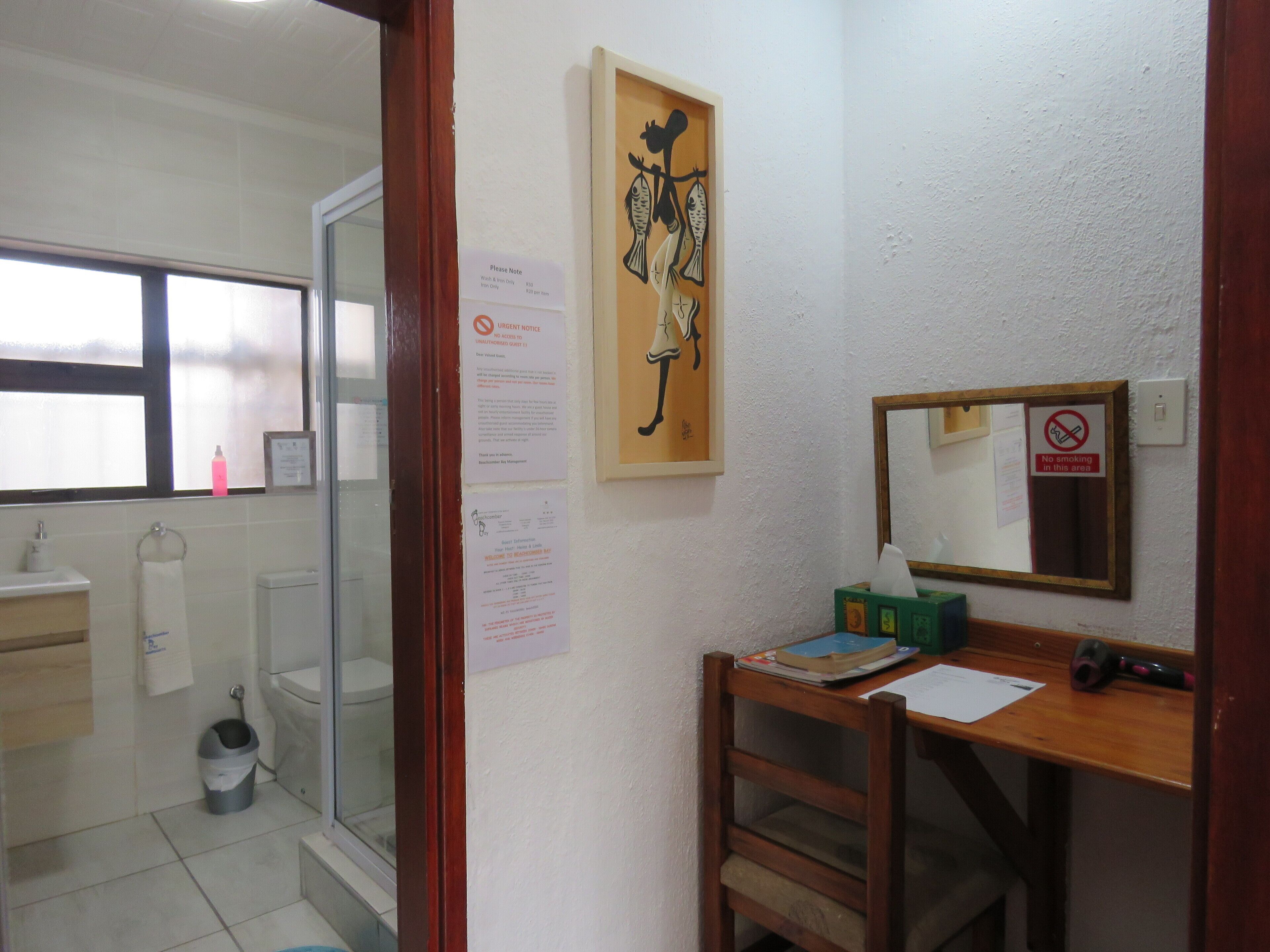 Una habitación pequeña y limpia con un escritorio de madera, un espejo y una ventana. Hay varios avisos y un cuadro en la pared.