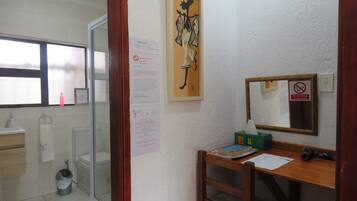 Una habitación pequeña y limpia con un escritorio de madera, un espejo y una ventana. Hay varios avisos y un cuadro en la pared.