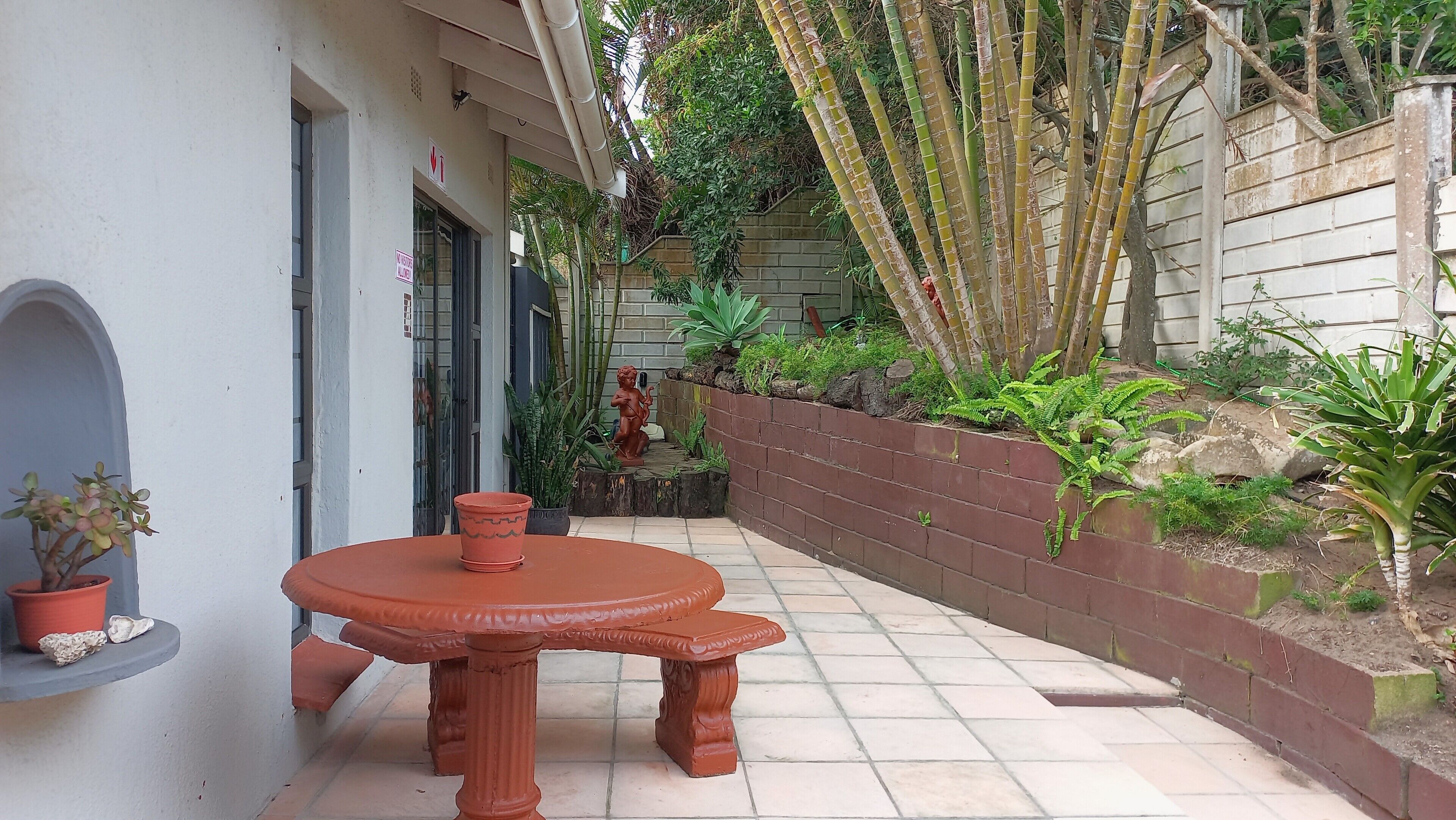 Eine Terrasse mit rundem Tisch und Stühlen, umgeben von einer Ziegelmauer mit Pflanzen und einer Statue.