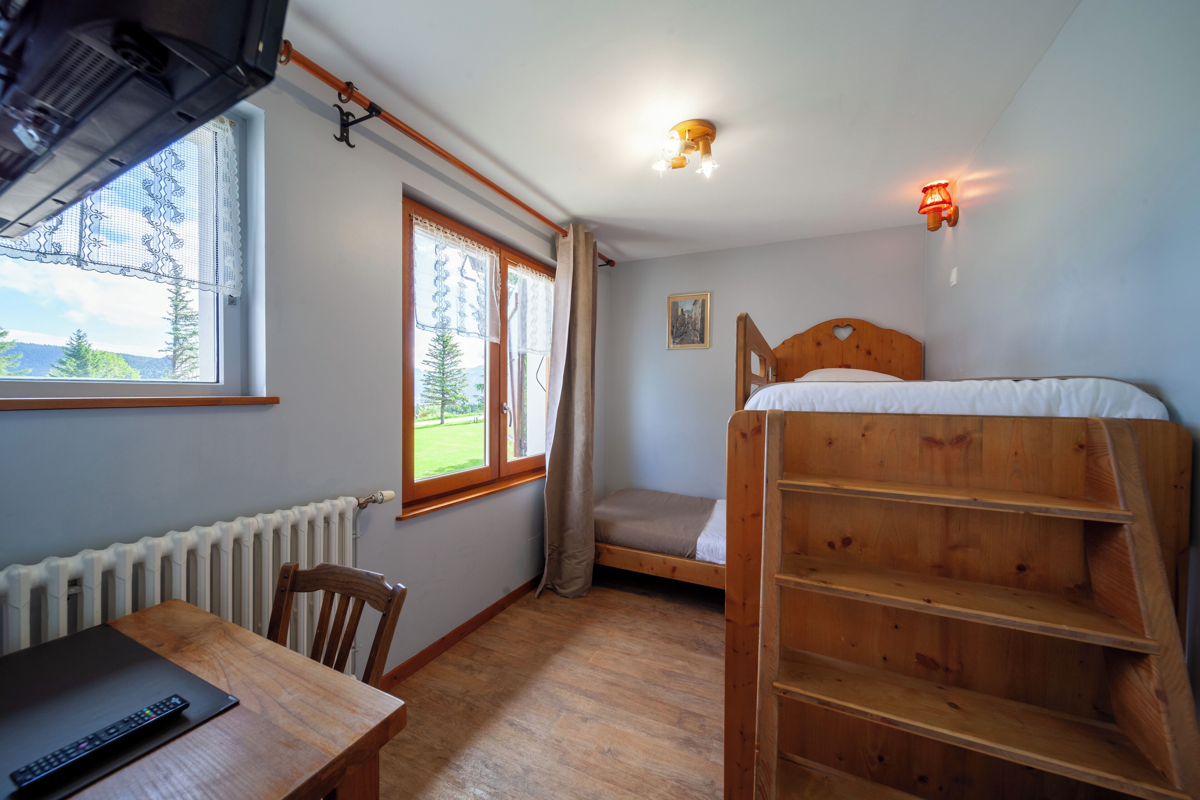 Una habitación con literas, un escritorio de madera con una computadora portátil, una silla, un radiador, una ventana con cortinas y una lámpara en el techo.