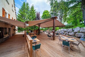 An outdoor patio with wooden flooring, tables, and chairs, surrounded by greenery and rocks. - Auberge des Montauds - Hôtel & Restaurant (Villard-de-Lans)