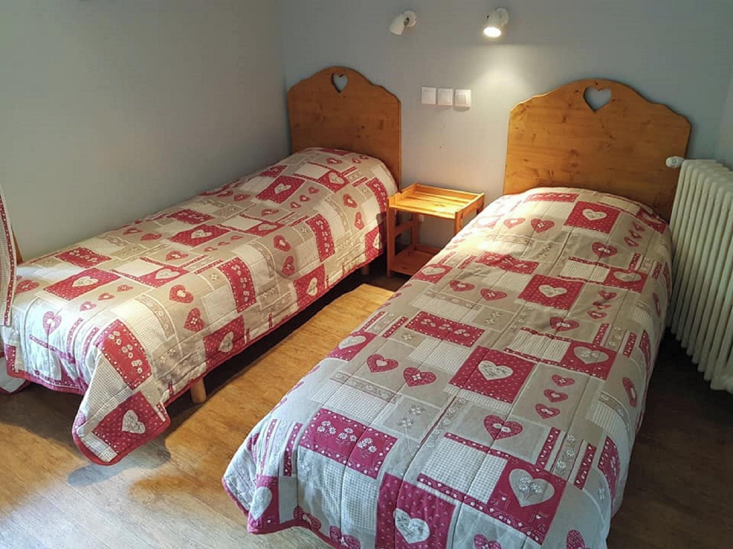 A bedroom with two single beds, each with a wooden headboard and a quilted bedspread featuring heart patterns. There is a small wooden nightstand between the beds, and a radiator is visible on the right wall.
