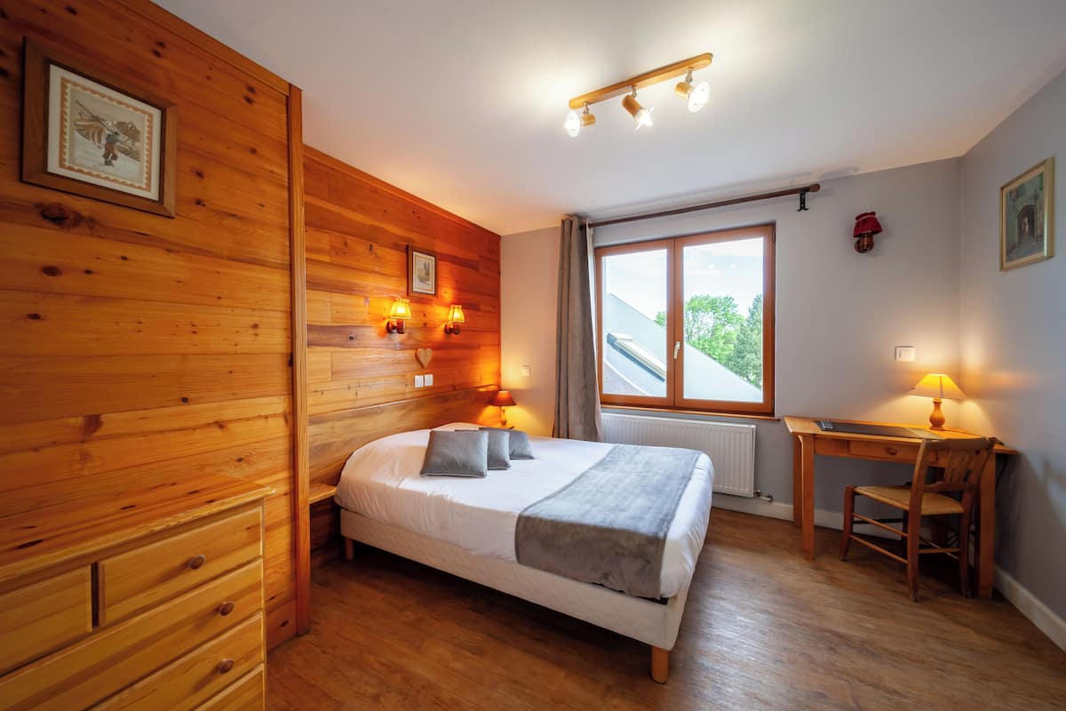 A wooden-paneled bedroom with a bed, bedside lamps, a desk, and a window with a view.