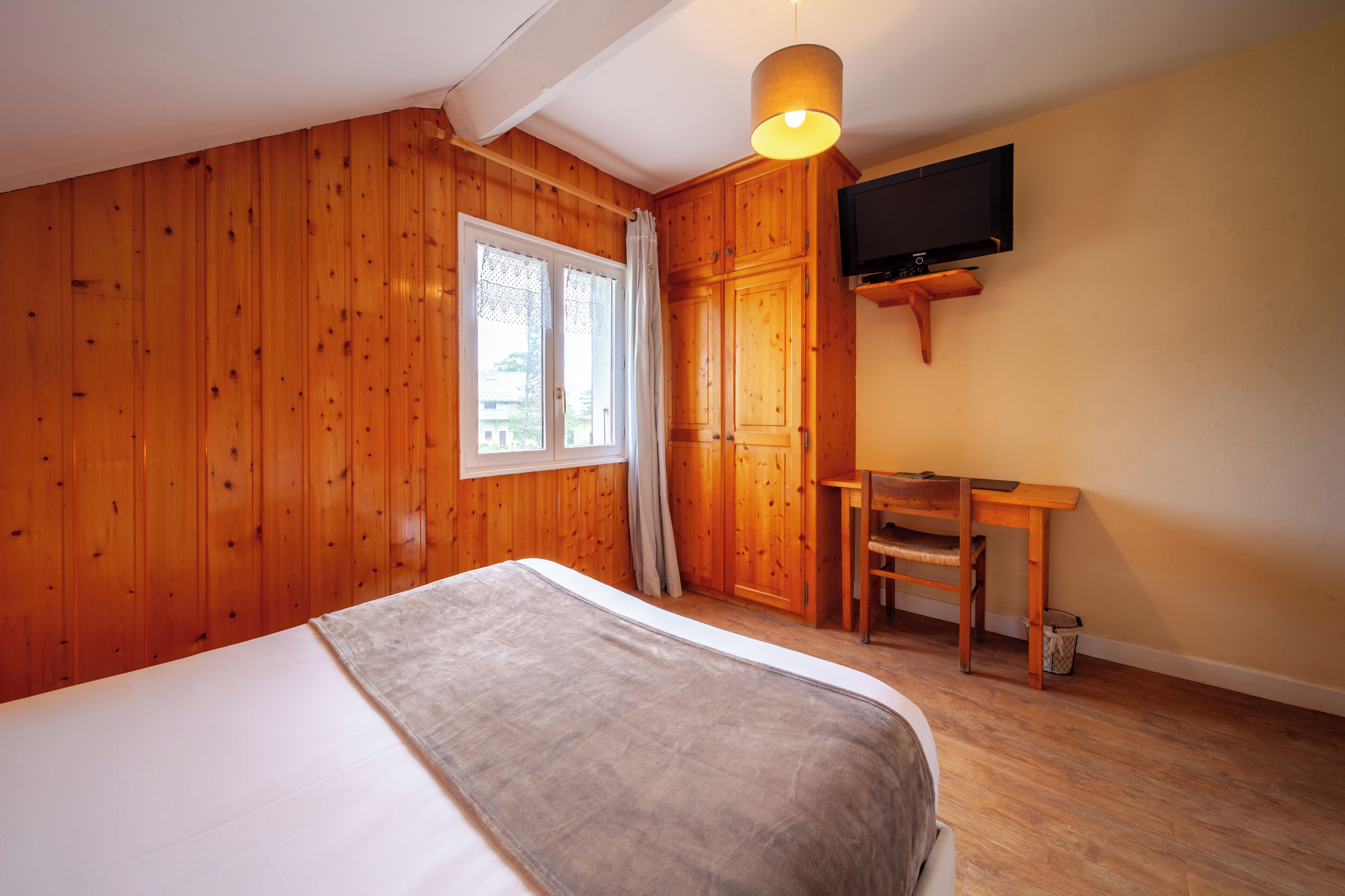 Une pièce aux murs en bois, un lit recouvert d’une couverture grise, un bureau avec une chaise et un téléviseur fixé au mur.