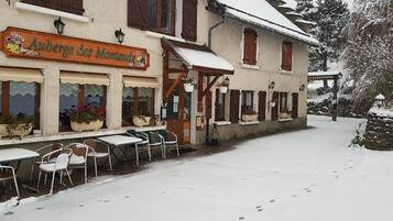 A snow-covered outdoor seating area with tables and chairs in front of a building with a sign reading "Auberge des Montaud."