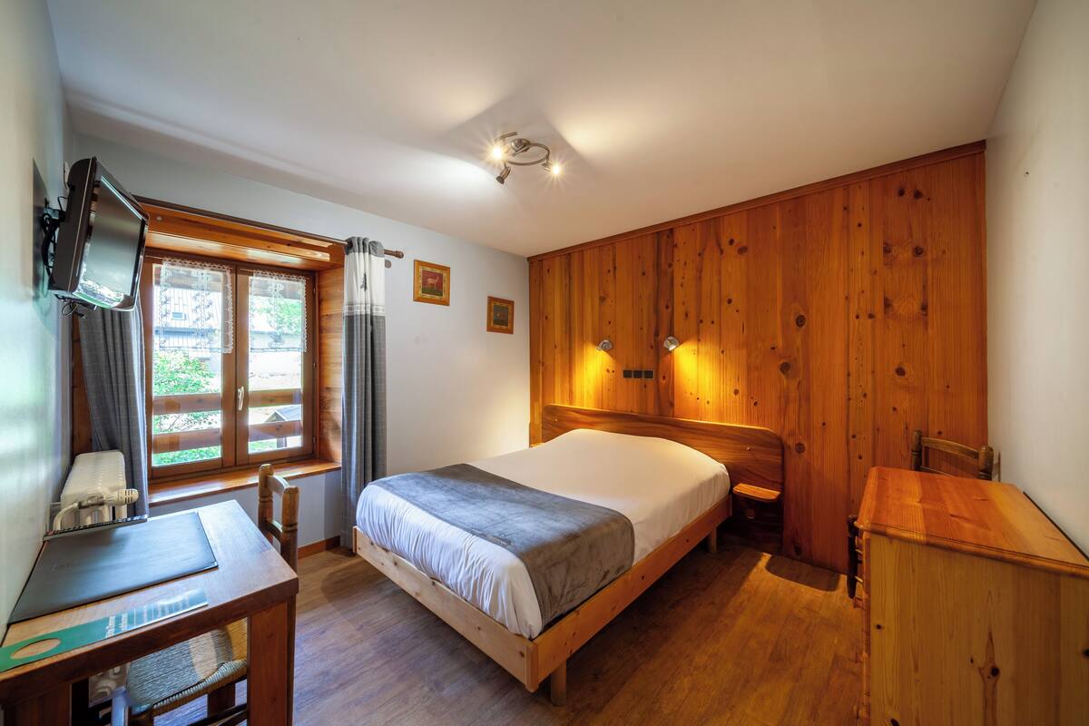 A bedroom with a wooden bed, a desk with a chair, a television, and a window with curtains.