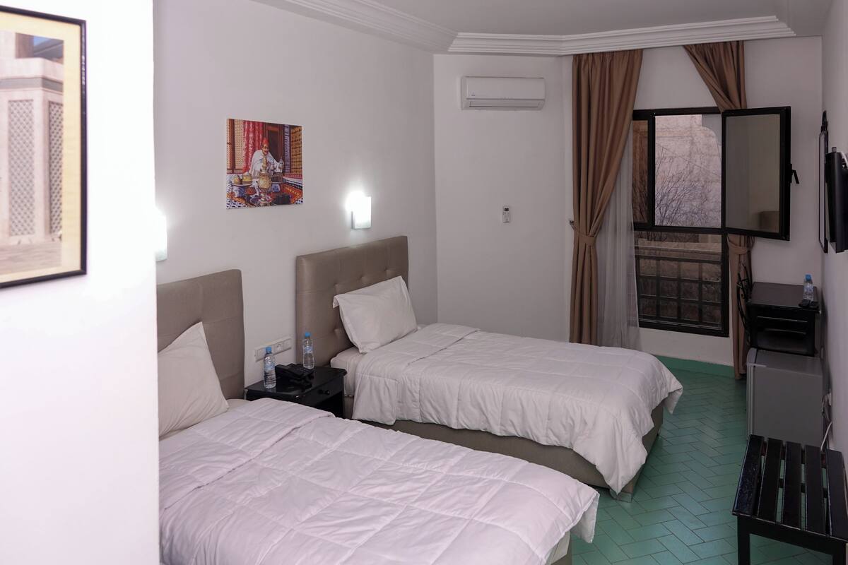 A hotel room with two beds, a TV, an air conditioning unit, a desk, and a window with curtains.