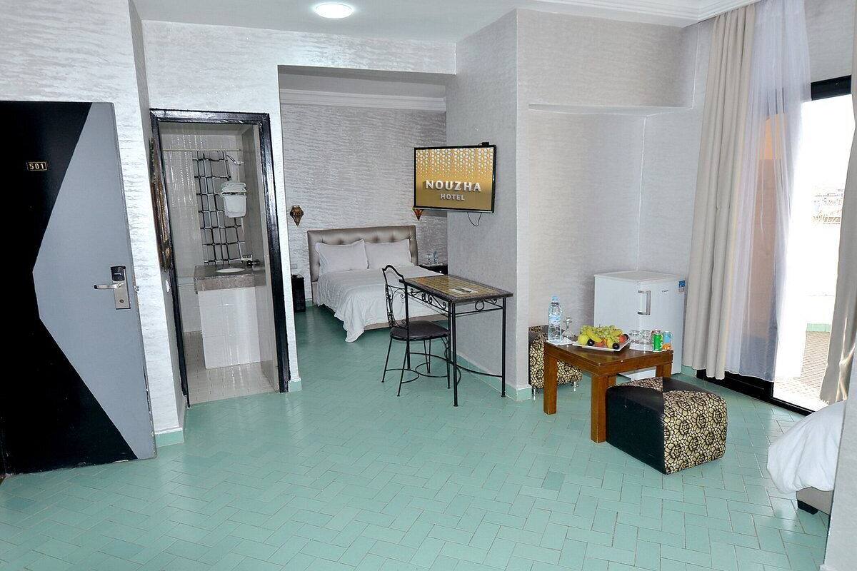 A hotel room with a bed, a desk, a television, and a small table with a bottle and fruit.
