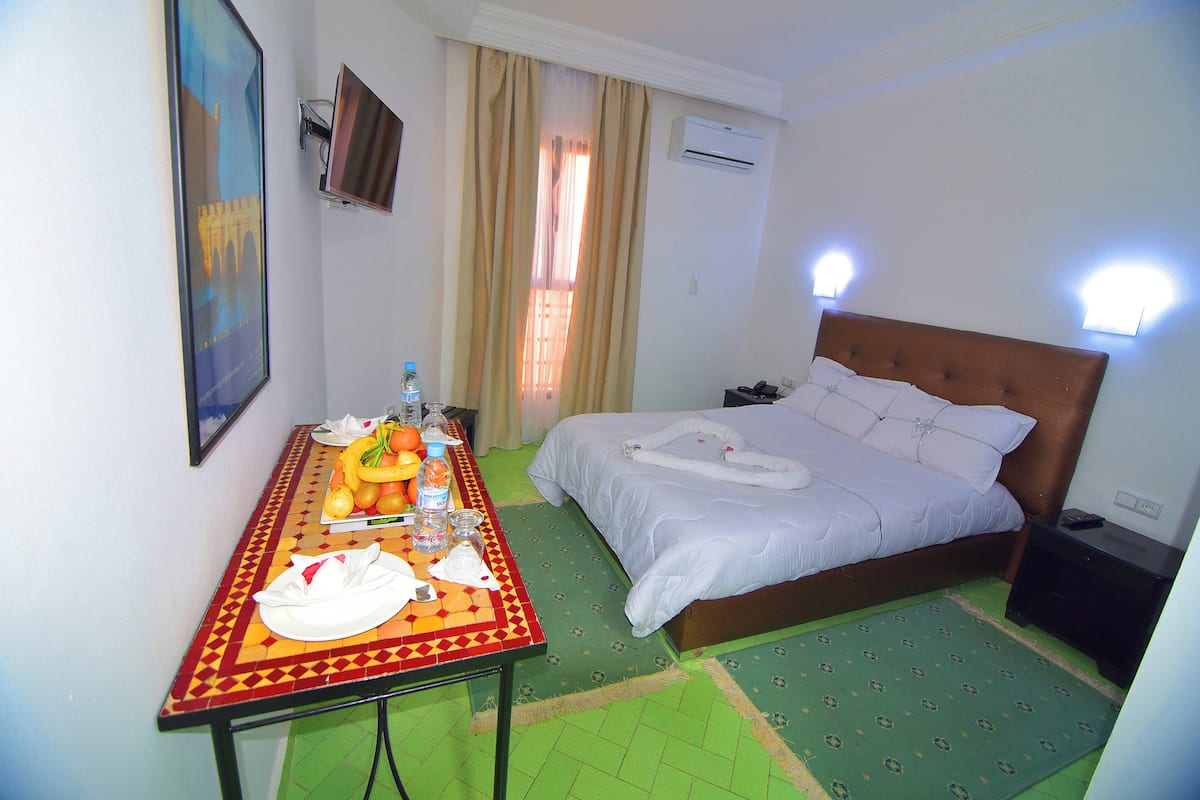 A hotel room with a bed, a table with breakfast items, a painting, and an air conditioning unit.